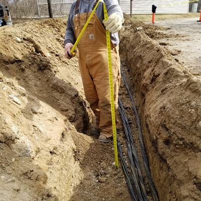 DisplayMax construction worker measuring trench depth for underground electrical conduit installation.